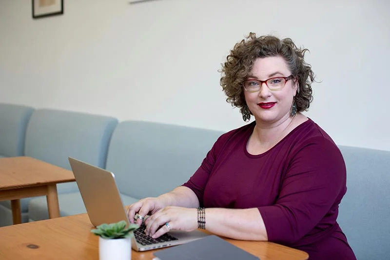 Lockige Frau mit Brille in einem roten Kleid sitzt in einem Café auf einem langen Sofa an einem Tisch und tippt auf ihrem Laptop.