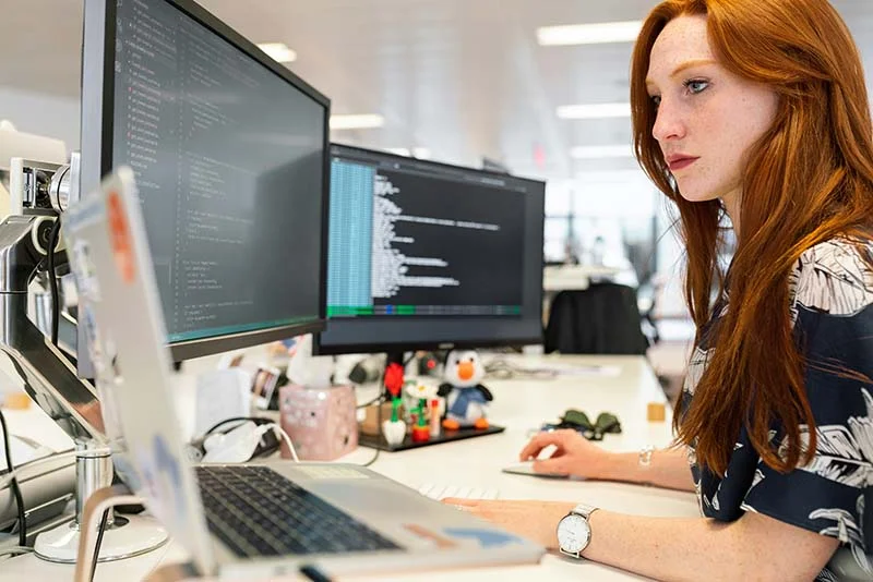 Junge Softwareentwicklung sitzt vor drei Bildschirmen mit Software-Code.